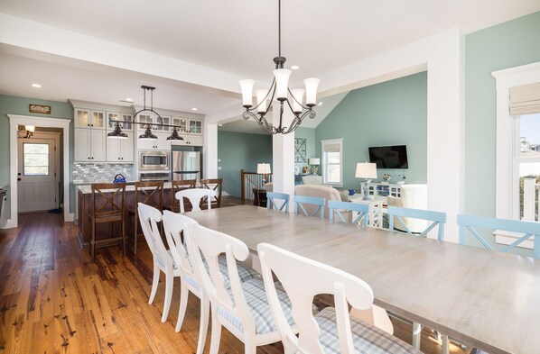 Una cocina amplia con suelo de madera, sillas blancas y azules alrededor de una mesa de comedor central, y una araña.