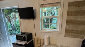 Cocina compacta con microondas, un refrigerador pequeño, un bote de basura, una silla de madera, una mesa, un televisor montado en la pared y una ventana con vistas a follaje.