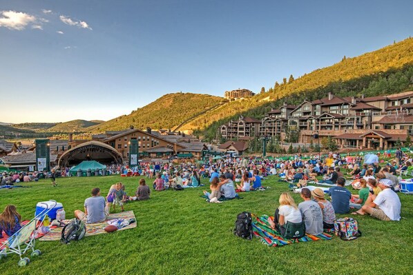 A large open grassy area with people sitting on blankets, a stage, and buildings in the background.