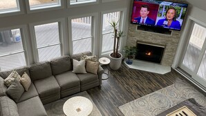 A modern living room with a sectional sofa, a fireplace, a television, and large windows offering a view of the outside. - 5-Star Oceanblock Sleeps 18! Luxury Beach Vacation Home Private Beach Community  (Bethany Beach)