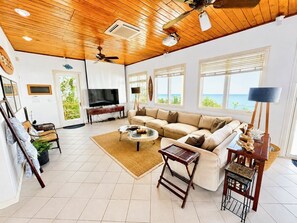 A spacious living room with a large sofa, a coffee table, and a TV. There are windows with a view of the sea, a ceiling fan, and a wooden ceiling.