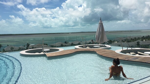 Ein Pool mit Holzterrasse und Liegestühlen, umgeben von einer Glasbarriere, mit Blick auf Strand und Meer.