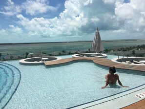 Una piscina con cubierta de madera y sillas de descanso, rodeada por una barrera de vidrio, con vista a una playa y al océano.