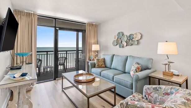 A modern living room with a sofa, coffee table, and a television. There is a balcony with a view of the ocean. - Luxurious Oceanfront Condo- Right on the Beach (Windy Hill)