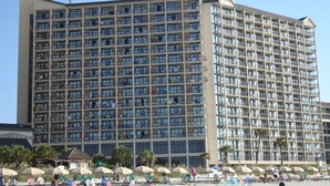 A large hotel with multiple floors and balconies, located near a beach with umbrellas and beach chairs. - Luxurious Oceanfront Condo- Right on the Beach (Windy Hill)