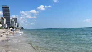 A beach with clear shallow waters, a sandy shore, and high-rise buildings in the background.