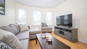 A living room with a sofa, ottoman, coffee table, TV, and windows.