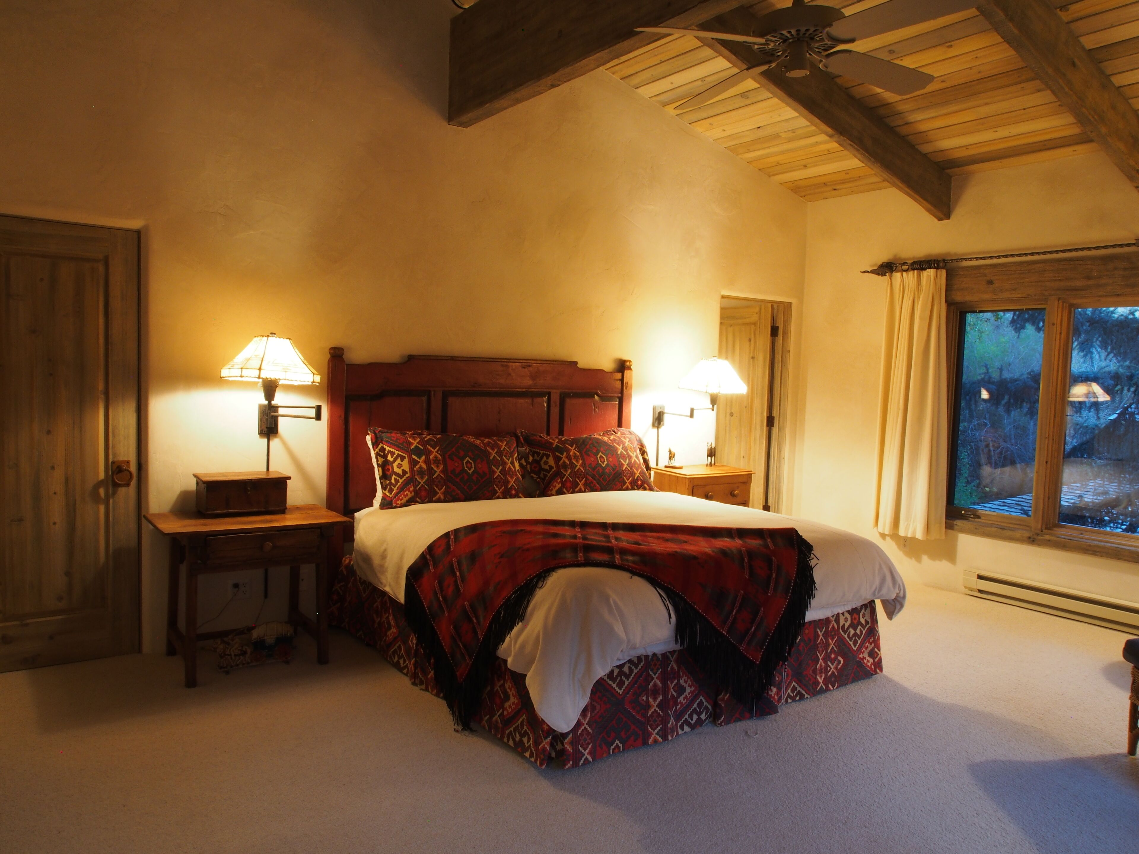 Un dormitorio con cabecera de madera, dos mesitas de noche con lámparas, una cama grande con ropa de cama estampada y una ventana con cortinas.