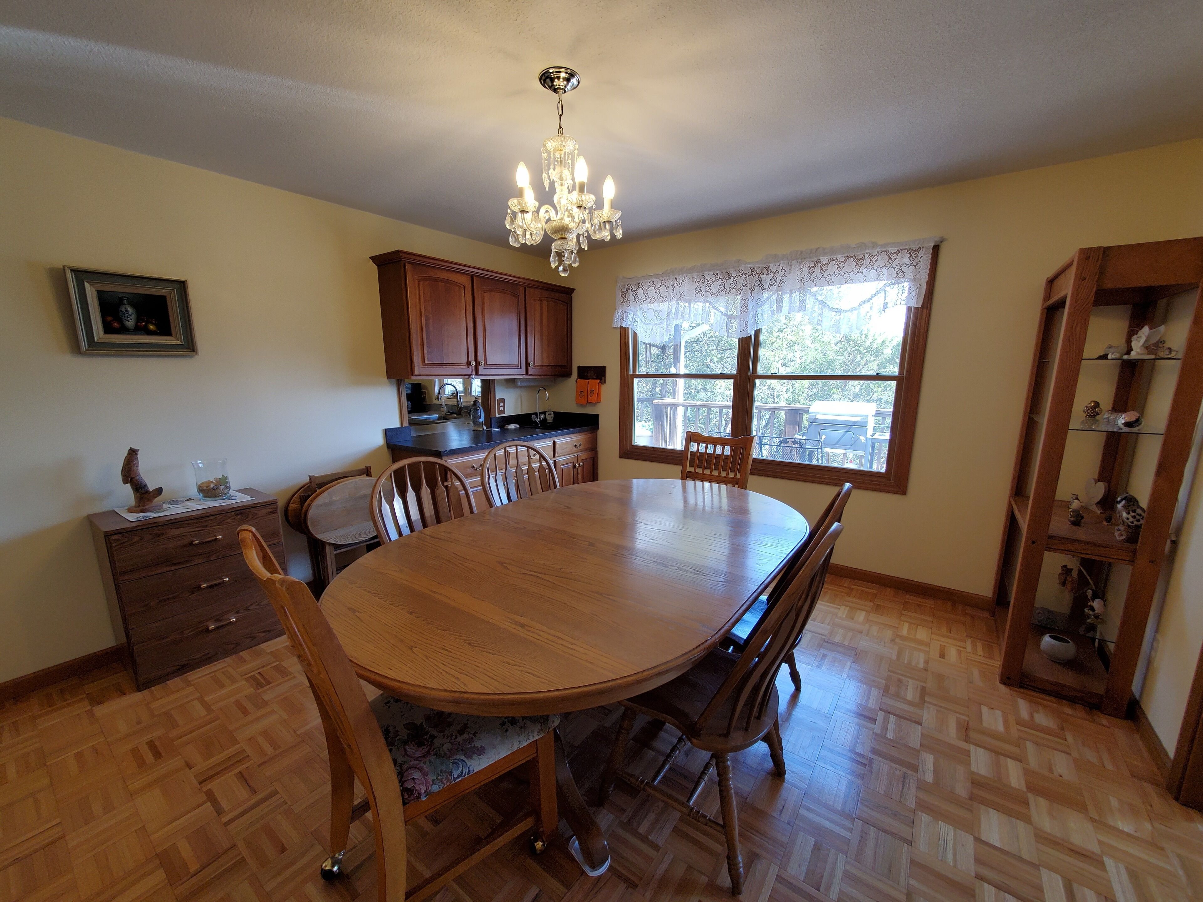 Un coin repas avec une table et des chaises en bois, un lustre, des meubles en bois et une fenêtre ornée de rideaux en dentelle.
