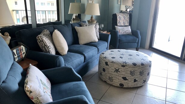 A living room with a blue sofa set, a round ottoman with nautical patterns, and a view of the city through large windows. - Beautiful Florida Condo in Oceanfront Building with River and Ocean Views (Daytona Beach Shores)