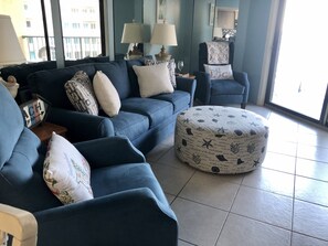 A living room with a blue sofa set, a round ottoman with nautical patterns, and a view of the city through large windows. - Beautiful Florida Condo in Oceanfront Building with River and Ocean Views (Daytona Beach Shores)