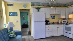 Cocina compacta con armarios blancos, refrigerador, microondas y estufa. Hay un sofá azul con una lámpara y un cuadro en la pared.