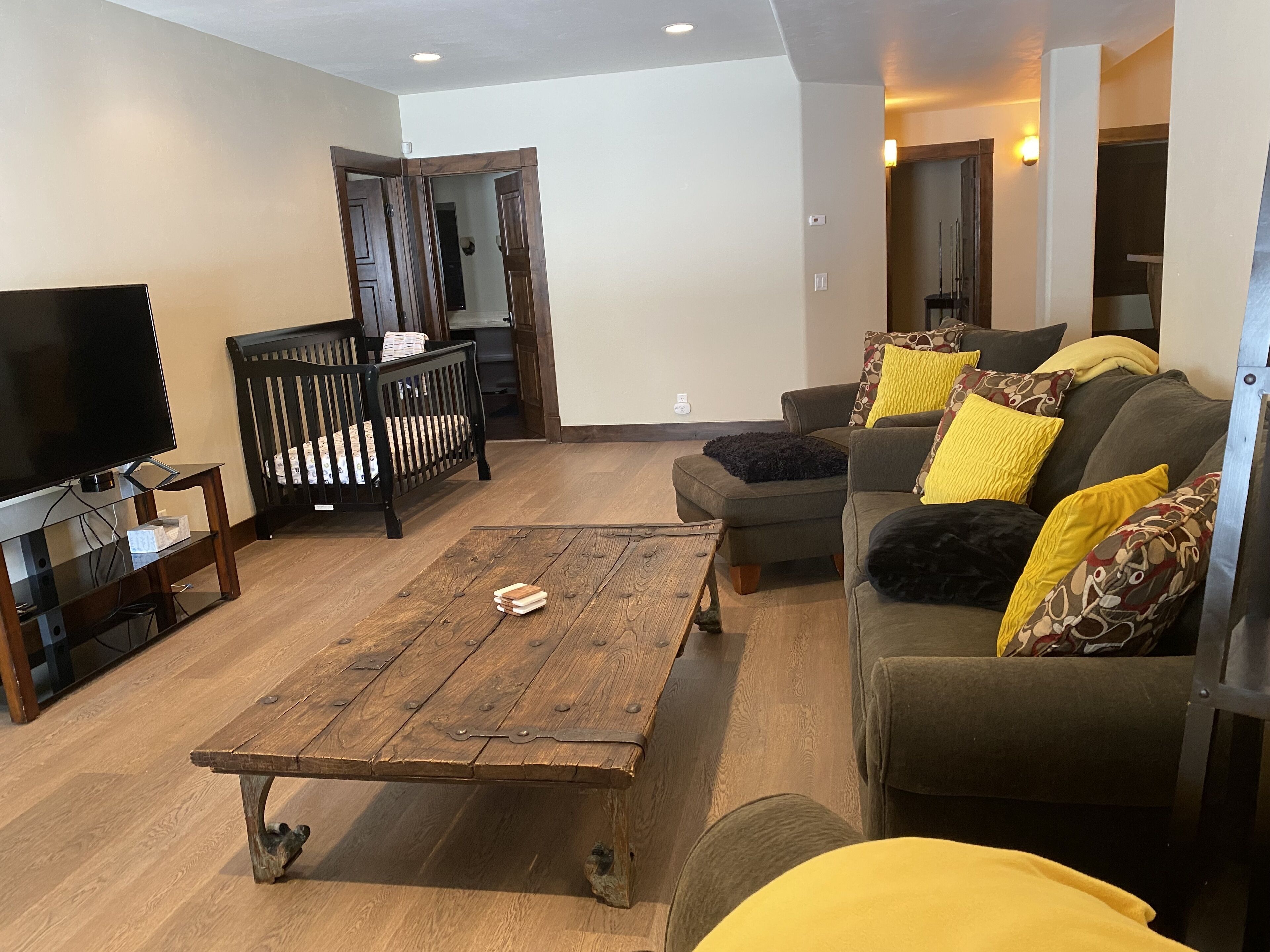 A living room with a wooden coffee table, a television, a crib, and a sofa with patterned cushions.