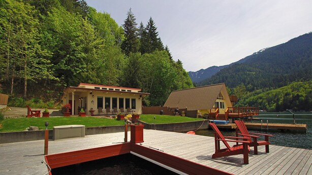 A wooden dock with red chairs, a small building with a red roof, and a lake surrounded by greenery.