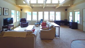 A spacious living room with a high ceiling, large windows, a flat-screen TV, leather sofas, a coffee table, and a desk with a computer.