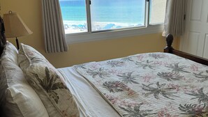 Une chambre à coucher avec un lit, des oreillers, une fenêtre avec des rideaux, une lampe et une vue sur l’océan.