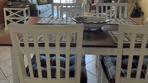 Un coin repas avec une table en bois et des chaises blanches, un canapé, une lampe et un téléviseur, le tout dans une pièce offrant une vue sur l’extérieur grâce à une porte coulissante.