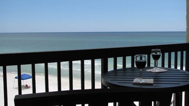 Un balcon avec une table et deux verres de vin rouge, donnant sur une plage où le ciel est d’un bleu limpide et la mer est calme.