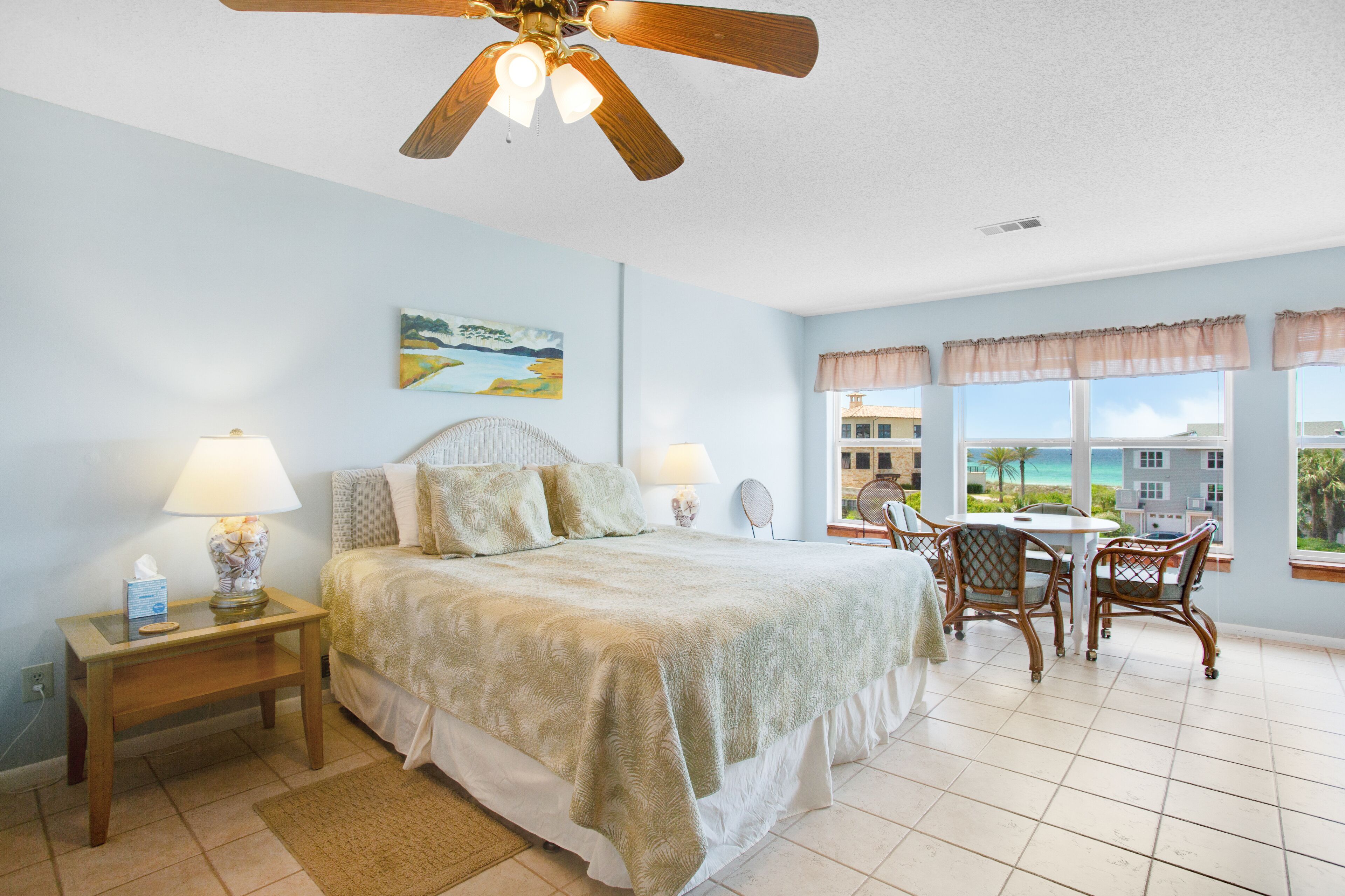 Une chambre à coucher comprenant un lit, des lampes de chevet, un ventilateur de plafond, une fenêtre donnant sur la plage et une petite table avec des chaises.