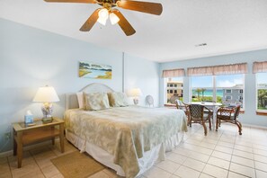 A bedroom with a bed, bedside lamps, a ceiling fan, a window with a view of the beach, and a small table with chairs. - Gulf View! Easy Beach and Pool Access. (Santa Rosa Beach)