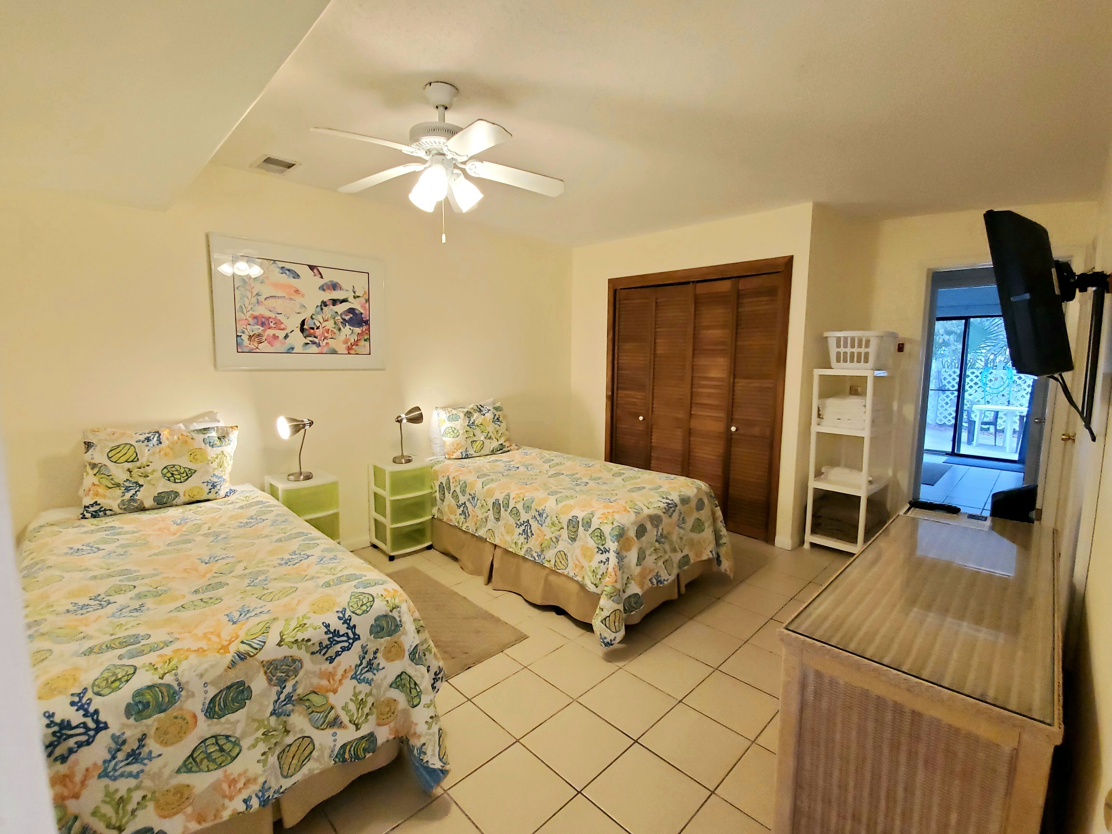 A bedroom with two beds, a ceiling fan, a wooden wardrobe, a TV, and a window with a view of the outside.