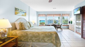 A bedroom with a bed, a desk, and a chair. There is a window with a view of the ocean.
