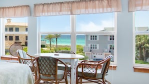 A room with a round table and wicker chairs, large windows with curtains, and a view of the beach and buildings.