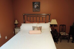 Una cama bien hecha con cabecera, una mesita de noche con lámpara, una silla y un cuadro colgado en la pared.