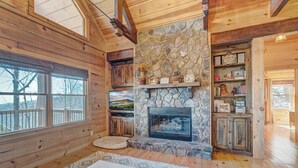 Interior acollidor d'una cabana amb una llar de pedra, parets de fusta i una finestra gran que dona a un paisatge nevós.
