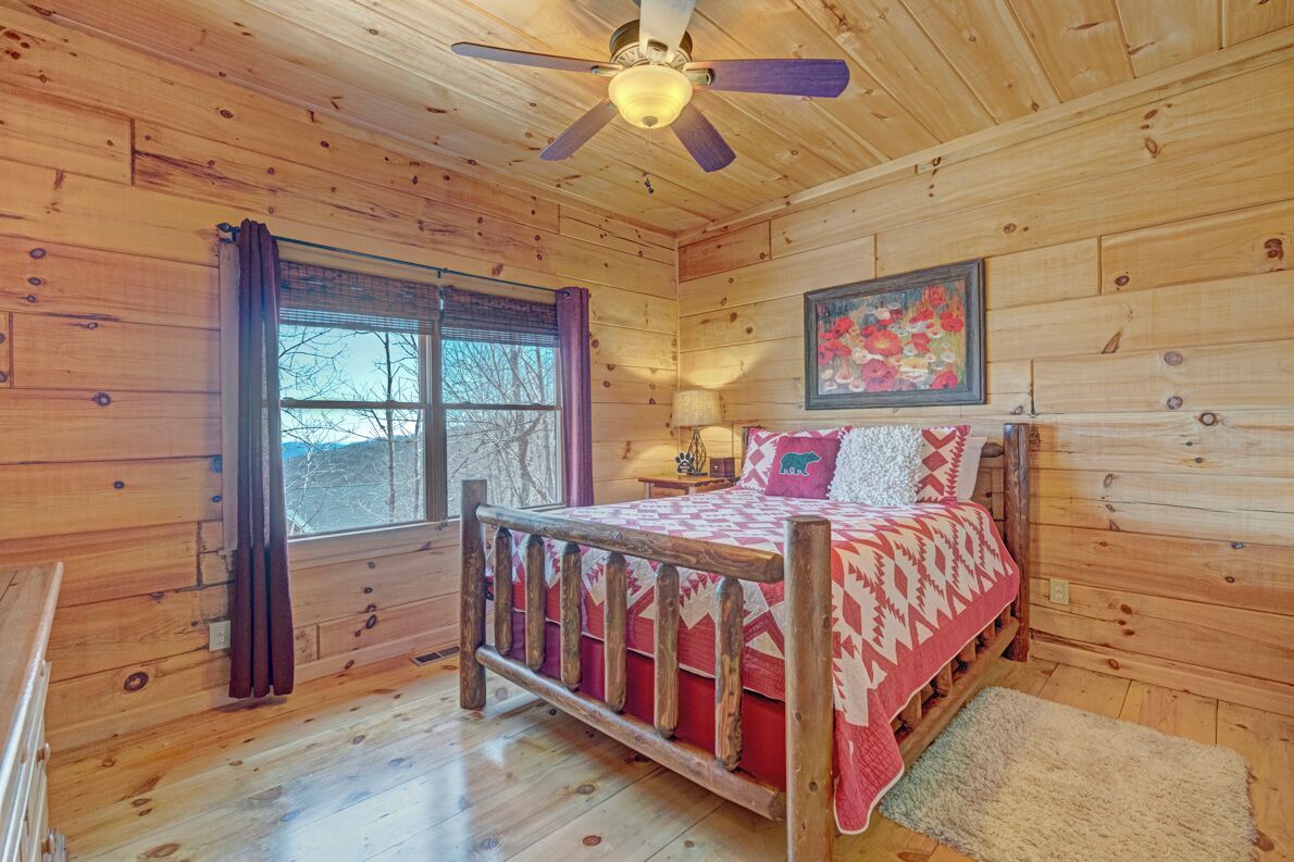 A wooden cabin bedroom with a bed, a ceiling fan, a window with curtains, and a painting on the wall.