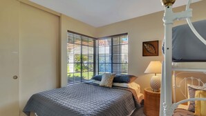 A bedroom with a bed, a nightstand, a lamp, a window with blinds, and a wall-mounted artwork.
