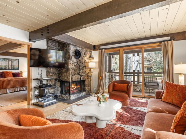 A cozy living room with a stone fireplace, comfortable seating, and a large window offering a view of the outdoors.