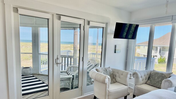 A room with a large sliding glass door leading to a balcony with a view of the beach. There is a white sofa and a bed with a white duvet.