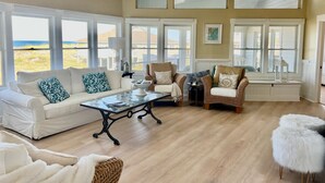 A spacious living room with a white sofa, wicker chairs, and a glass coffee table. Large windows offer a view of the beach.