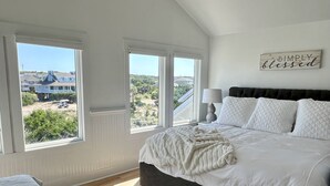 A bedroom with a large bed, a bedside table with a lamp, and a view of houses through the windows.