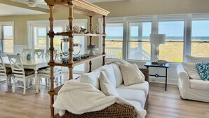 A spacious living room with a high ceiling, large windows offering a view of the beach, a wooden bookshelf, and a cozy seating area with a white sofa and a wicker chair.