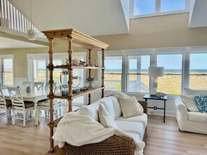 Relax in this bright and airy living area with a stunning beach view, featuring a comfortable sofa, wicker chair, and large windows.