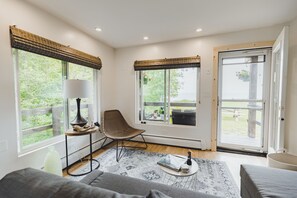 A modern living room with a sofa, a chair, a small table with a lamp, and large windows with blinds. - Charming LAKE SUPERIOR Beach Cottage in Keweenaw, near Calumet. NORTHERN LIGHTS! (Calumet)