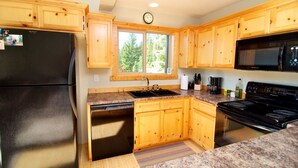 Une cuisine avec des meubles en bois, un réfrigérateur noir, une cuisinière, un four à micro-ondes et une fenêtre donnant sur des arbres.