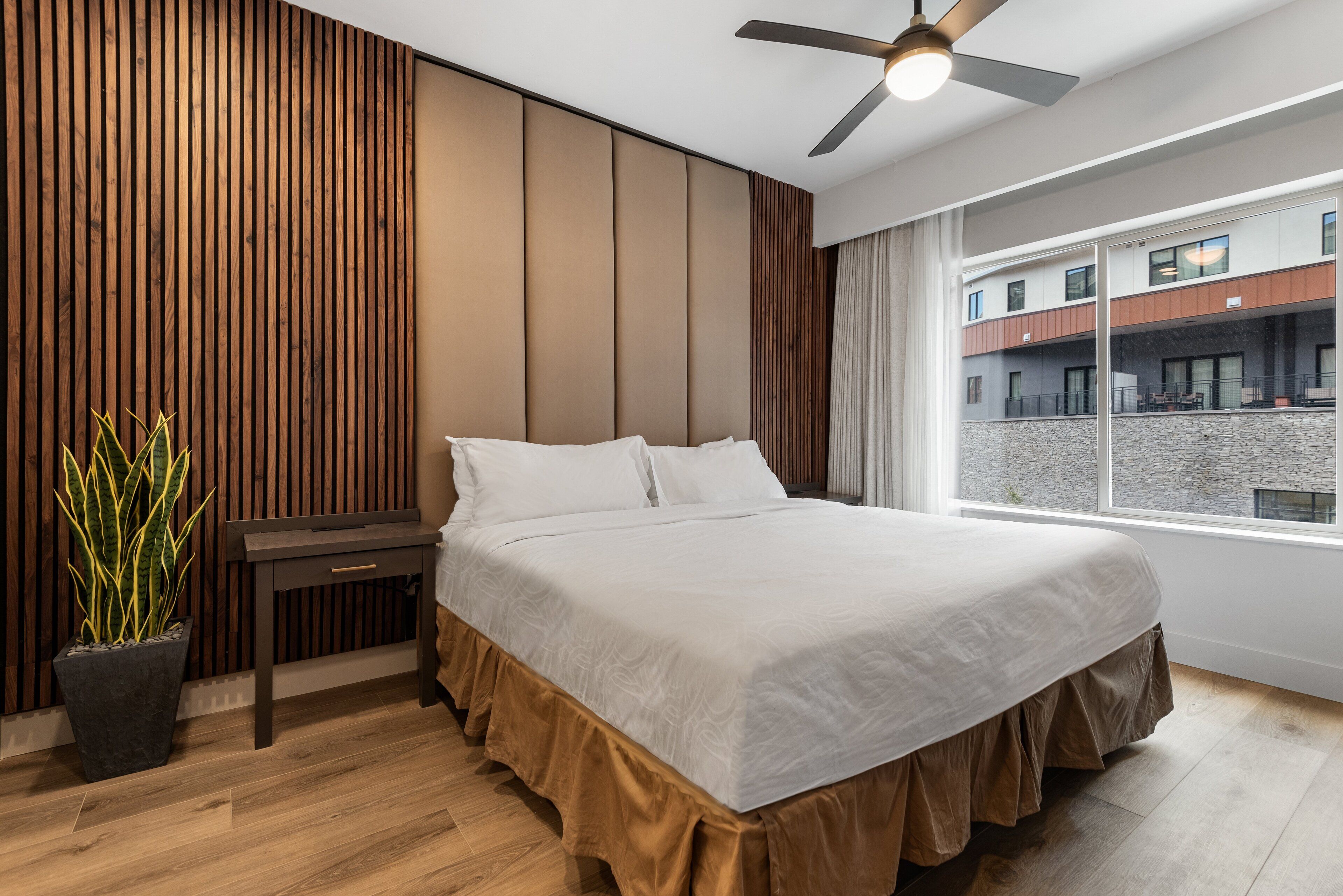 Ein modernes Schlafzimmer mit einem großen Bett, einem Deckenventilator, einer Topfpflanze und Blick auf das Gebäude draußen.