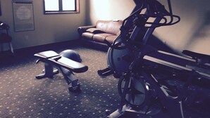 Een sportschool thuis met trainingsapparatuur, een raam, een bank en een aan de muur bevestigde televisie.