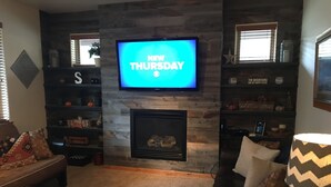 Een woonkamer met een open haard, een televisie waarop 'NEW THURSDAY' staat, en een gezellige zithoek met een bank en voetenbankje.