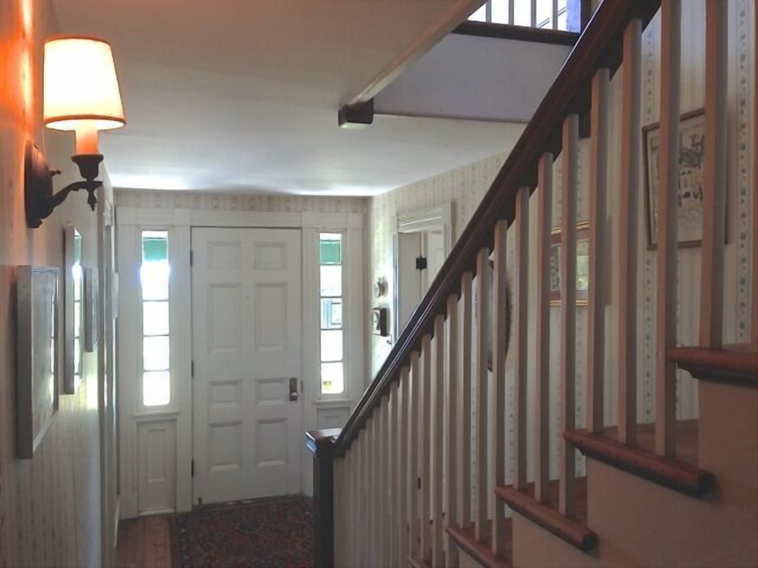 A hallway with a wooden staircase, a white door, and a wall-mounted lamp.