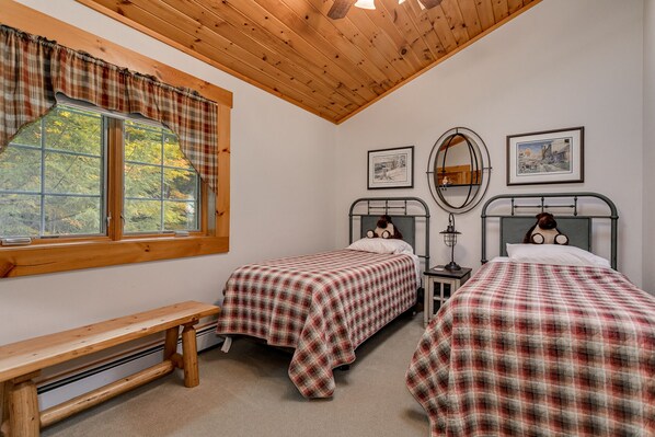 A bedroom with two beds, a bench, a window with curtains, and framed pictures on the wall.