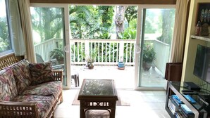 A living room with a sofa, a coffee table, and a TV. There is a sliding glass door leading to a balcony with plants and a wooden fence. - Spacious, Comfortable with Full Kitchen & W/D (Princeville)