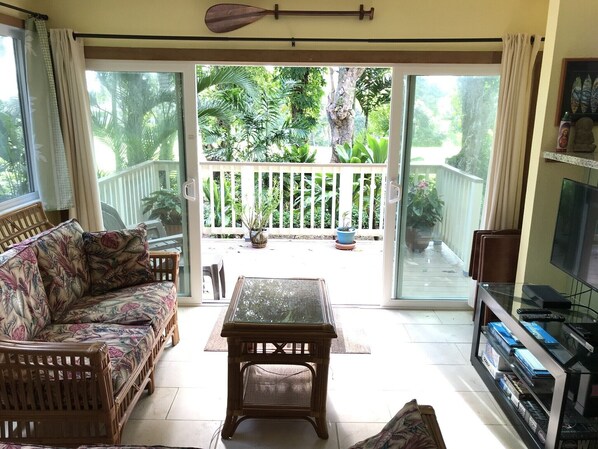 A living room with a sofa, a coffee table, and a TV. There is a sliding glass door leading to a balcony with plants and a wooden fence. - Spacious, Comfortable with Full Kitchen & W/D (Princeville)