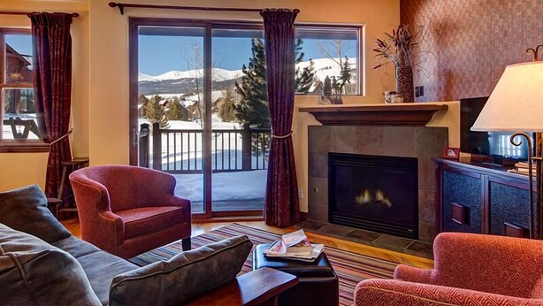 A cozy living room with a fireplace, a sofa, a chair, and a view of snow-capped mountains through large windows. - Breckenridge 3 Bedrm Townhouse, Private Hot Tub, near Golf, Hiking & Bike trails (Breckenridge)