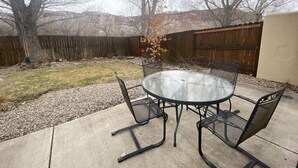 Een terras met een ronde glazen tafel en vier metalen stoelen, omringd door een houten hek en een boom.