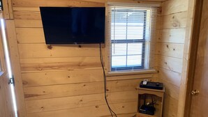 Quarto com painéis de madeira, TV montada na parede, janela com persianas e um armário pequeno com aparelhos eletrônicos.