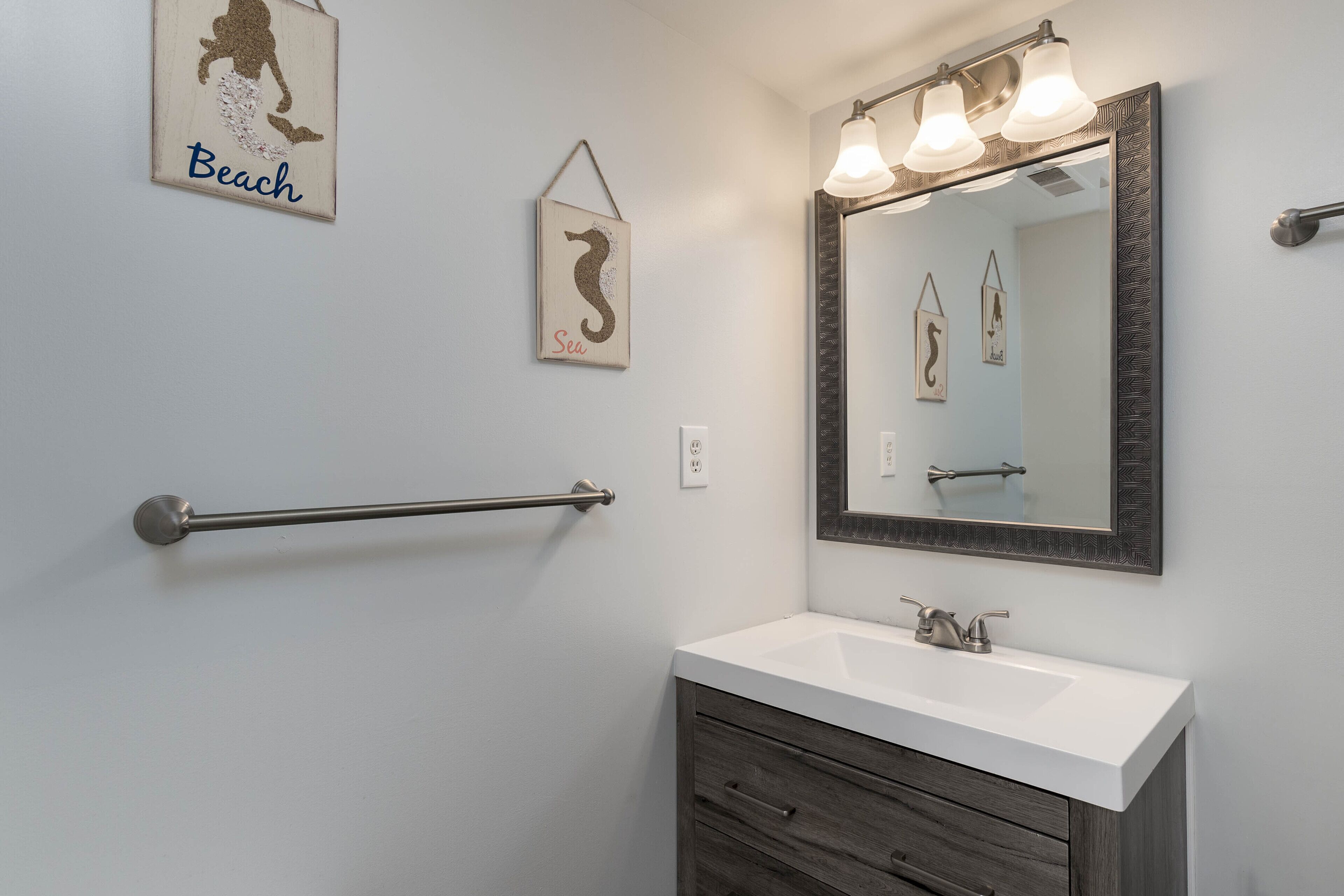A bathroom with a white sink, a large mirror with lights, a towel rack, and wall art.